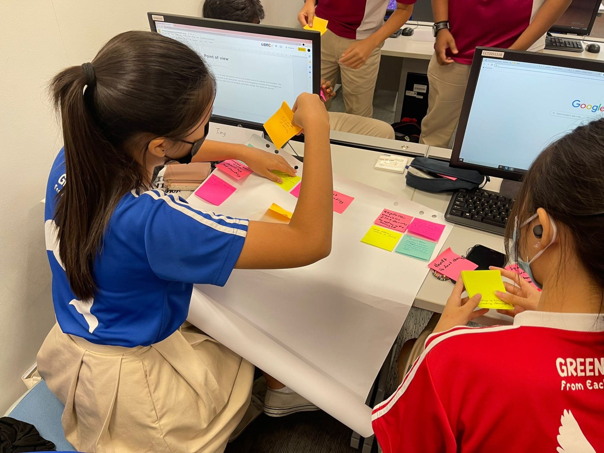 Two students placing colorful sticky notes on a white sheet at a desk with computer monitors nearby.